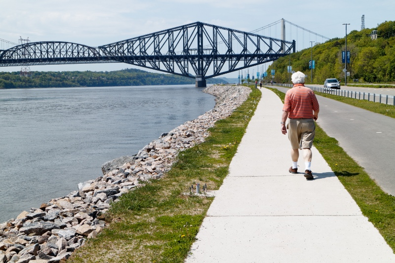 An older man walking on Promenade Samuel de Champlain park in Quebec City, with the St Lawrence river at Pont du Quebec bridge beyond