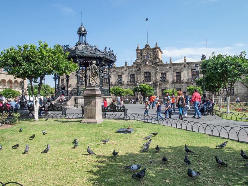 Scene of urban life in Plaza de Armas in historic center, people walking, pigeons on grass, kiosk and Government building in background, sunny day