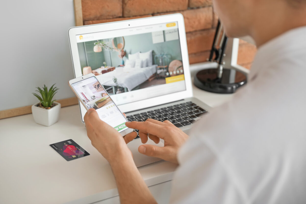 Man looking and booking a hotel room online using phone and a laptop