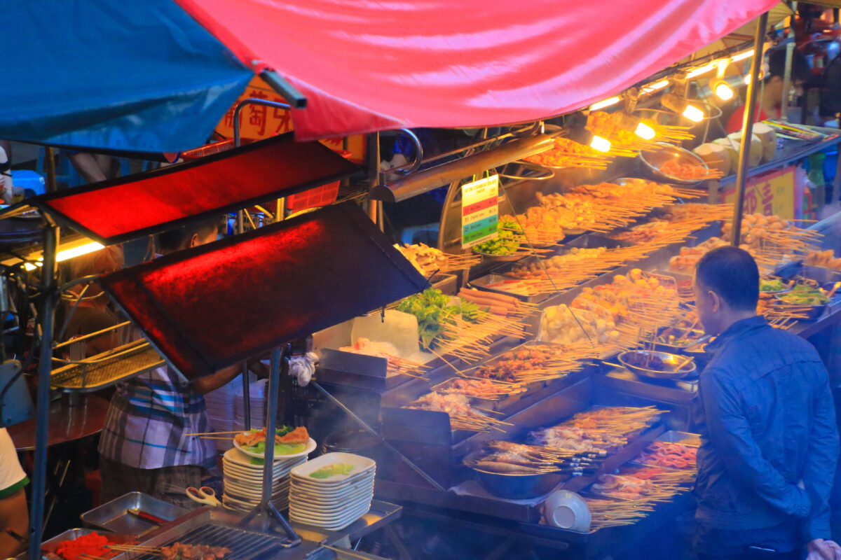 Man standing in front a street food stall selling a huge variety of food in skewers