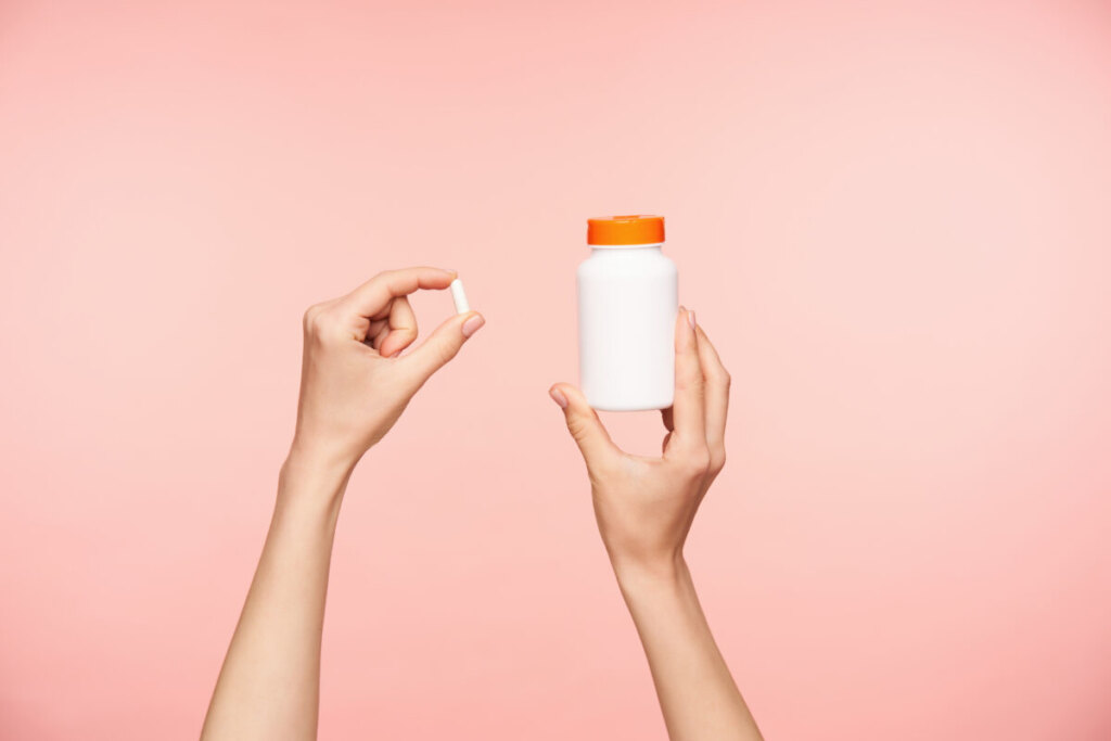 Hands holing a white pill and white bottle with orange cover of vitamins